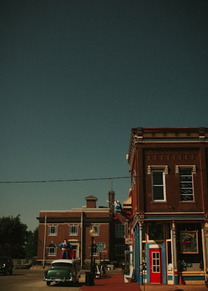 A vintage-style street scene features brick buildings with ornate trim. Statues resembling iconic superheroes are displayed on the building and street, adding a whimsical touch. A classic car is parked in front, enhancing the retro atmosphere. The sky is clear and the overall scene evokes a sense of nostalgia.