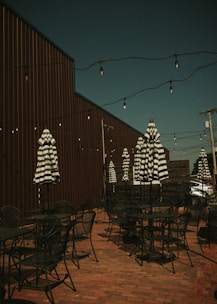 An outdoor patio area features several black metal tables and chairs, with striped black and white umbrellas providing potential shade. The area is decorated with hanging string lights above, and a tall wooden fence runs alongside the space. The patio is set against a brick floor, creating a rustic and cozy ambiance.