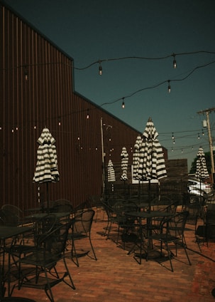 An outdoor patio area features several black metal tables and chairs, with striped black and white umbrellas providing potential shade. The area is decorated with hanging string lights above, and a tall wooden fence runs alongside the space. The patio is set against a brick floor, creating a rustic and cozy ambiance.