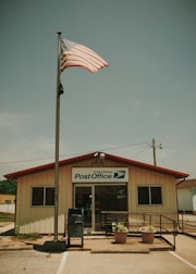 A small, single-story post office building with a beige exterior and a red roof stands under a blue sky. An American flag waves atop a tall flagpole in front of the building. Large windows flank the entrance, and there is a blue mailbox outside. Two potted plants with flowers sit by the entrance, and a ramp with railings leads to the door.