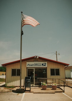 A small, single-story post office building with a beige exterior and a red roof stands under a blue sky. An American flag waves atop a tall flagpole in front of the building. Large windows flank the entrance, and there is a blue mailbox outside. Two potted plants with flowers sit by the entrance, and a ramp with railings leads to the door.