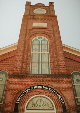 A tall, red-brick church tower with arched windows featuring stained glass designs. The lower part of the tower displays the inscription 'The Master is here and calls for you' above a circular stained glass window. The architectural style is classic, with a symmetrical facade and decorative brickwork.