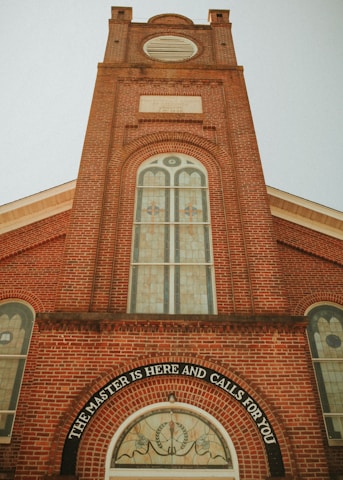 A tall, red-brick church tower with arched windows featuring stained glass designs. The lower part of the tower displays the inscription 'The Master is here and calls for you' above a circular stained glass window. The architectural style is classic, with a symmetrical facade and decorative brickwork.