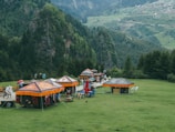 Several colorful tents with ethnic patterns are set up on a grassy area surrounded by lush green mountains. People are walking around the tents, some with umbrellas. The background consists of dense forests and distant rolling hills with scattered villages.
