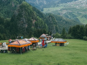 Several colorful tents with ethnic patterns are set up on a grassy area surrounded by lush green mountains. People are walking around the tents, some with umbrellas. The background consists of dense forests and distant rolling hills with scattered villages.