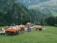 Several colorful tents with ethnic patterns are set up on a grassy area surrounded by lush green mountains. People are walking around the tents, some with umbrellas. The background consists of dense forests and distant rolling hills with scattered villages.