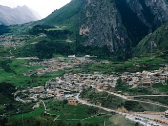 A scenic view of a large village nestled among lush green hills and towering mountains. The village is composed of clustered buildings with traditional architectural styles, surrounded by greenery and terraced fields. A winding road leads through the village, with vehicles parked along it. The background features rugged mountains with steep cliffs and dense forests.