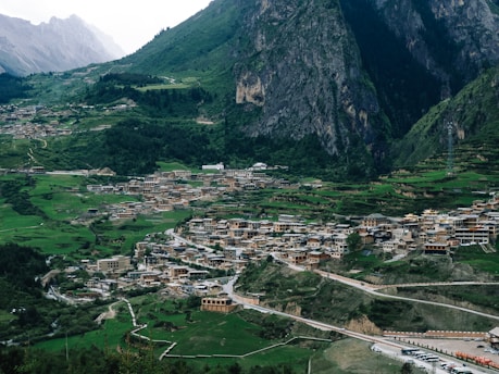 A scenic view of a large village nestled among lush green hills and towering mountains. The village is composed of clustered buildings with traditional architectural styles, surrounded by greenery and terraced fields. A winding road leads through the village, with vehicles parked along it. The background features rugged mountains with steep cliffs and dense forests.
