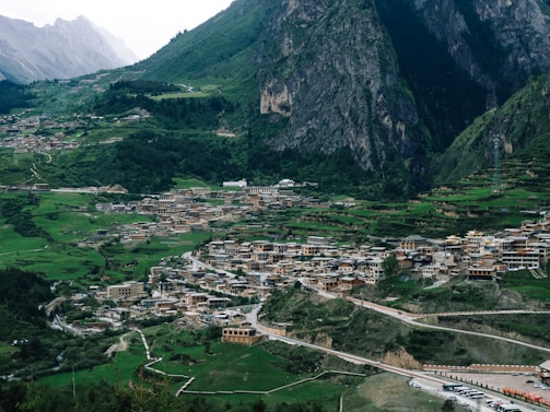 A scenic view of a large village nestled among lush green hills and towering mountains. The village is composed of clustered buildings with traditional architectural styles, surrounded by greenery and terraced fields. A winding road leads through the village, with vehicles parked along it. The background features rugged mountains with steep cliffs and dense forests.