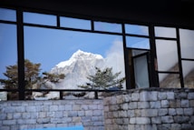 A snow-capped mountain peak is visible through the large glass window of a building. The reflection includes trees and the clear blue sky. The building features stone walls and the window design creates a frame-like effect around the natural scene.