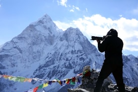 A person holding a camera with a large lens is positioned on a rocky outcrop, capturing images of a majestic snow-covered mountain peak. Colorful prayer flags are strung across the foreground, adding a vibrant contrast to the snow and rocks. The sky is mostly clear with some scattered clouds, enhancing the backdrop.