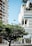 Modern high-rise buildings with architecture featuring glass facades and greenery on balconies. A large tree with dense leaves is in the foreground, with blue skies above. The setting appears to be urban, with streetlights and paved pathways.