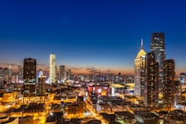 A vibrant cityscape captured at dusk, featuring numerous skyscrapers illuminated against a deep blue and orange sky. The buildings are modern and varied in design, with bright lights highlighting the urban landscape. Streets and buildings are lit, creating a lively and bustling atmosphere.