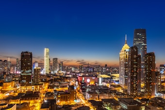 A vibrant cityscape captured at dusk, featuring numerous skyscrapers illuminated against a deep blue and orange sky. The buildings are modern and varied in design, with bright lights highlighting the urban landscape. Streets and buildings are lit, creating a lively and bustling atmosphere.