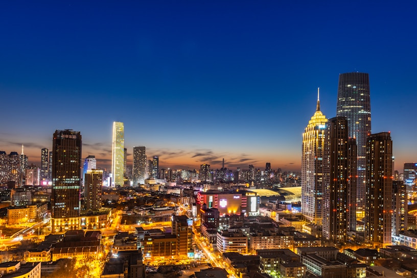 A vibrant cityscape captured at dusk, featuring numerous skyscrapers illuminated against a deep blue and orange sky. The buildings are modern and varied in design, with bright lights highlighting the urban landscape. Streets and buildings are lit, creating a lively and bustling atmosphere.