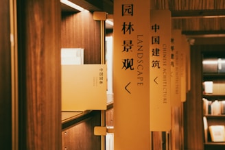 A series of bookshelves lined with books are labeled with vertical signs. The focus is on a sign with the words 'LANDSCAPE' in English and Chinese characters. The lighting is warm, creating a cozy atmosphere in the wooden interior.