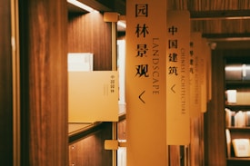 A series of bookshelves lined with books are labeled with vertical signs. The focus is on a sign with the words 'LANDSCAPE' in English and Chinese characters. The lighting is warm, creating a cozy atmosphere in the wooden interior.