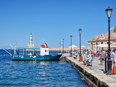 A glass-bottom boat named Stella is docked at a marina, with people walking along the busy promenade. In the background, a lighthouse stands on a stone pier, and there are several umbrella-shaded tables near historic-looking domed buildings. The sky is clear, and the sea is calm, reflecting the vibrant blue of the sky.