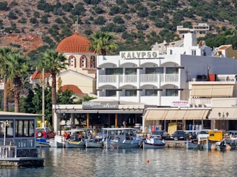 A picturesque coastal town with a mix of traditional and modern architecture. A white building with a large balcony and the name Kalypso is prominently displayed. In the foreground, several small boats are docked along the waterfront, with palm trees and a church with a red dome in the background.