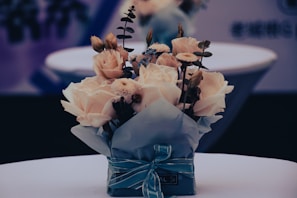 A bouquet of flowers neatly arranged and wrapped in blue paper with a ribbon. The bouquet includes roses and other small flowers, set against a soft background with neutral tones.
