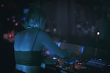 A person with short hair is engaged in DJing at a dimly lit venue. The individual is standing at a turntable with various controls and illuminated displays. Soft ambient lighting casts a blue hue over the person and the equipment.