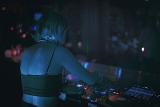 A person with short hair is engaged in DJing at a dimly lit venue. The individual is standing at a turntable with various controls and illuminated displays. Soft ambient lighting casts a blue hue over the person and the equipment.