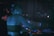 A person with short hair is engaged in DJing at a dimly lit venue. The individual is standing at a turntable with various controls and illuminated displays. Soft ambient lighting casts a blue hue over the person and the equipment.
