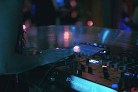 A DJ booth is dimly lit with a variety of glowing lights on the equipment. A person is using the sound mixer, with their hand poised over the controls. The scene is full of electronic equipment with a focus on a Pioneer DJ controller.