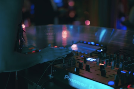 A DJ booth is dimly lit with a variety of glowing lights on the equipment. A person is using the sound mixer, with their hand poised over the controls. The scene is full of electronic equipment with a focus on a Pioneer DJ controller.