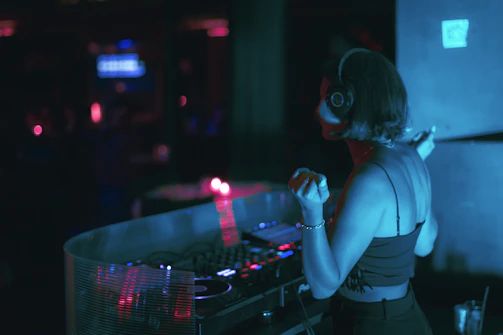 A DJ wearing headphones and a black sleeveless top is standing at a DJ booth, illuminated by blue and pink lights. The scene suggests a dimly lit club atmosphere, with equipment visible on the booth. A soft focus blurs the background, contributing to the intimate, dynamic setting of a music venue.