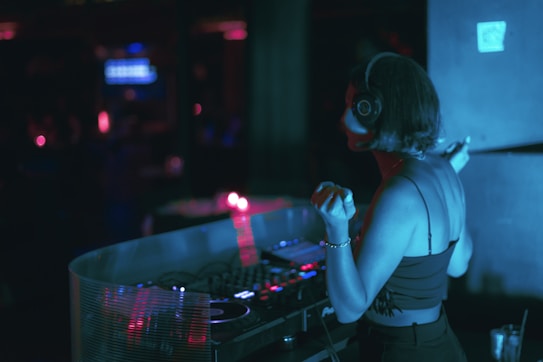 A DJ wearing headphones and a black sleeveless top is standing at a DJ booth, illuminated by blue and pink lights. The scene suggests a dimly lit club atmosphere, with equipment visible on the booth. A soft focus blurs the background, contributing to the intimate, dynamic setting of a music venue.