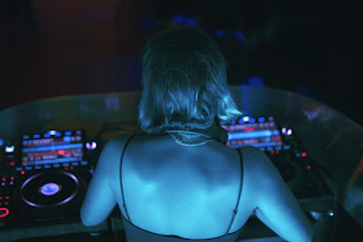 A person with short hair wearing headphones stands in front of DJ equipment. The lighting is dim with a blue hue, and the focus is on their back as they engage with the turntables and mixer.