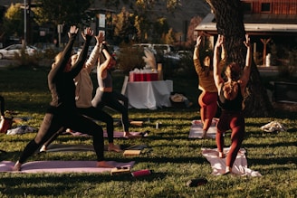 A group of people engaged in an outdoor yoga session on a grassy area. Six individuals are lined up on yoga mats executing a standing pose with arms raised towards the sky. The scene is lit by warm sunlight, creating a calm and focused atmosphere. In the background, there are trees, a set of tables covered with a white cloth, and a building across the street.