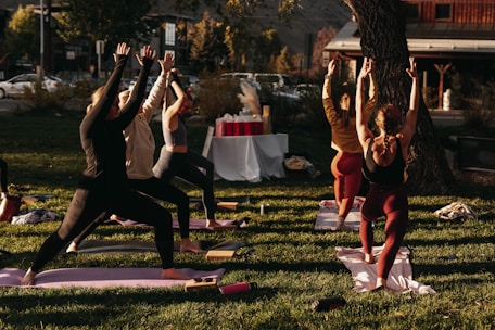 A group of people engaged in an outdoor yoga session on a grassy area. Six individuals are lined up on yoga mats executing a standing pose with arms raised towards the sky. The scene is lit by warm sunlight, creating a calm and focused atmosphere. In the background, there are trees, a set of tables covered with a white cloth, and a building across the street.