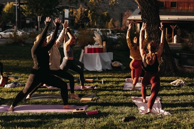 A group of people engaged in an outdoor yoga session on a grassy area. Six individuals are lined up on yoga mats executing a standing pose with arms raised towards the sky. The scene is lit by warm sunlight, creating a calm and focused atmosphere. In the background, there are trees, a set of tables covered with a white cloth, and a building across the street.