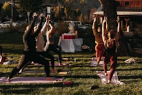 A group of people engaged in an outdoor yoga session on a grassy area. Six individuals are lined up on yoga mats executing a standing pose with arms raised towards the sky. The scene is lit by warm sunlight, creating a calm and focused atmosphere. In the background, there are trees, a set of tables covered with a white cloth, and a building across the street.