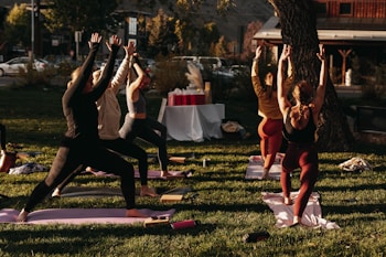 A group of people engaged in an outdoor yoga session on a grassy area. Six individuals are lined up on yoga mats executing a standing pose with arms raised towards the sky. The scene is lit by warm sunlight, creating a calm and focused atmosphere. In the background, there are trees, a set of tables covered with a white cloth, and a building across the street.