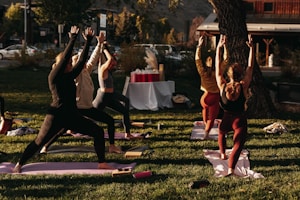 A group of people engaged in an outdoor yoga session on a grassy area. Six individuals are lined up on yoga mats executing a standing pose with arms raised towards the sky. The scene is lit by warm sunlight, creating a calm and focused atmosphere. In the background, there are trees, a set of tables covered with a white cloth, and a building across the street.
