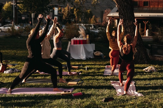 A group of people engaged in an outdoor yoga session on a grassy area. Six individuals are lined up on yoga mats executing a standing pose with arms raised towards the sky. The scene is lit by warm sunlight, creating a calm and focused atmosphere. In the background, there are trees, a set of tables covered with a white cloth, and a building across the street.