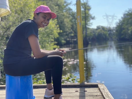 A person is sitting on a blue stool near a body of water, holding a fishing rod. The individual is wearing a pink cap, black t-shirt, and leggings, smiling while facing the water. The surroundings are lush with greenery, and there is a tranquil river reflecting the blue sky and trees.