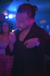 A person is using a lighter to ignite a cigarette in a dimly lit, atmospheric setting. The individual wears a black outfit and a stylish wristwatch, with their hair tied back. The background includes blurred figures, suggesting a social or nightlife environment with vibrant colored lights, primarily blue and purple.
