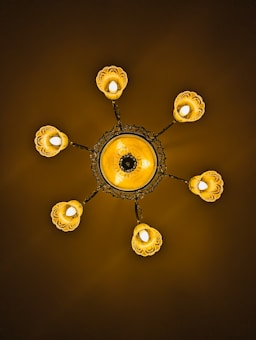 An ornate chandelier featuring a central circular design with intricate patterns. Six light fixtures extend outward, each enclosed in a petal-like covering. The lighting casts a warm, golden glow against a dark background.