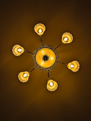 An ornate chandelier featuring a central circular design with intricate patterns. Six light fixtures extend outward, each enclosed in a petal-like covering. The lighting casts a warm, golden glow against a dark background.