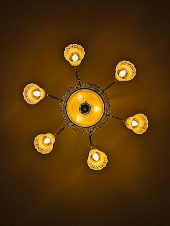 An ornate chandelier featuring a central circular design with intricate patterns. Six light fixtures extend outward, each enclosed in a petal-like covering. The lighting casts a warm, golden glow against a dark background.