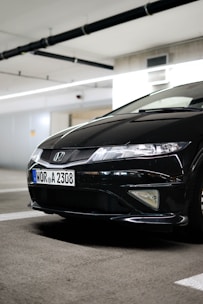 A black Honda car is parked in an indoor parking structure with visible license plate details. The environment is well-lit, and the car's front end, including its grille and headlights, are prominently displayed.