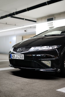 A black Honda car is parked in an indoor parking structure with visible license plate details. The environment is well-lit, and the car's front end, including its grille and headlights, are prominently displayed.