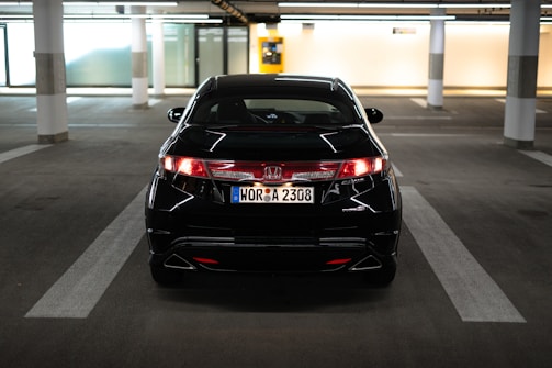 A black Honda car is parked in an indoor parking garage with white pillars and marked parking spaces. The license plate is visible, and the lighting is soft, with yellowish tones reflecting off the car's surface.