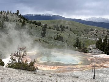 A hot spring is situated amidst a rugged landscape with gentle hills and sparse trees. The steam rising from the geothermal feature creates a mystical atmosphere. The colors of the spring range from blue and green in the center to orange and brown at the edges, contrasting with the surrounding terrain. In the background, there are distant mountains partially covered with snow under a cloudy sky.