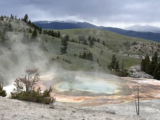 A hot spring is situated amidst a rugged landscape with gentle hills and sparse trees. The steam rising from the geothermal feature creates a mystical atmosphere. The colors of the spring range from blue and green in the center to orange and brown at the edges, contrasting with the surrounding terrain. In the background, there are distant mountains partially covered with snow under a cloudy sky.