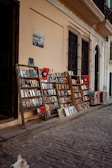 A narrow cobblestone street is lined with wooden racks displaying a variety of books and magazines. The racks are positioned against a light beige wall, with barred windows above them. A sign and posters add character to the scene. A small portion of a dog can be seen in the bottom left corner, enhancing the street market atmosphere.